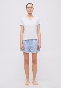 Γυναίκα που στέκεται ξυπόλυτη, φορώντας λευκό μπλουζάκι με κοντά μανίκια και V λαιμόκοψη και σορτς με μπλε και λευκές ρίγες, μπροστά σε απλό φόντο.