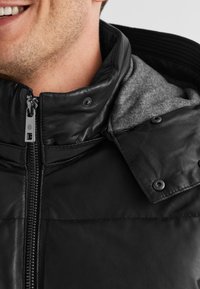 Schwarze Lederjacke mit einem grauen Innenfutter, ausgestattet mit einem Reißverschluss, Druckknöpfen und einem gerippten Kragen. Glatte Textur.