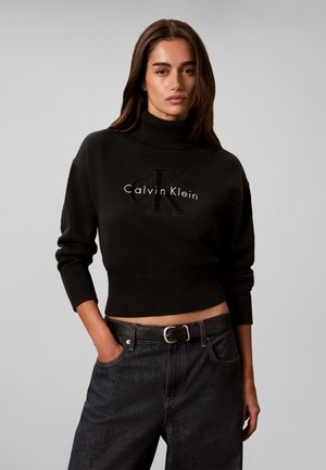 Kvinde iført en sort Calvin Klein cropped rullekrave sweater og mørke jeans med sort bælte, stående med den ene hånd i lommen.