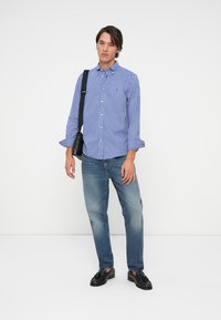 Niebieska i biała w kratę koszula z guzikami, dżinsowe jeansy i czarne buty. Model pozuje z czarną torbą. Prosty, casualowy design i fason.