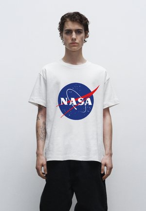 Bílý bavlněný tričko se modrým logem NASA včetně bílého textu, červeného švihu a hvězdných akcentů na přední straně. Model nosí černé kalhoty.