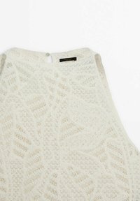 Haut blanc sans manches avec motif en dentelle florale texturée, fermeture par boucle et bouton au niveau de l'encolure arrière, et étiquette de marque visible à l'intérieur.