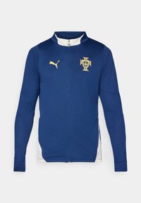 Giacca a zip blu realizzata in tessuto testurizzato con dettagli bianchi. Presenta un logo Puma dorato e lo stemma nazionale del Portogallo sul petto.