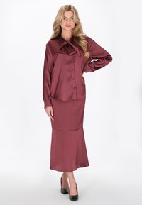 Bordó szatén ruha masnis gallérral, gombos díszítéssel, hosszú ujjakkal és bővülő szoknyarésszel, fekete magassarkú cipővel párosítva.