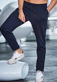 Marineblauwe joggers met een trekkoord in de taille en zijzakken, gemaakt van een zachte stof. Gecombineerd met witte sneakers met groene accenten.
