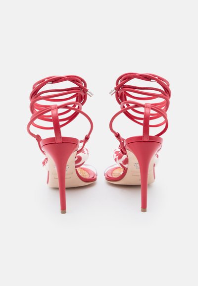 Sandales à talons hauts rouges avec des talons aiguilles fins et plusieurs brides à la cheville, vues de dos sur un fond clair.