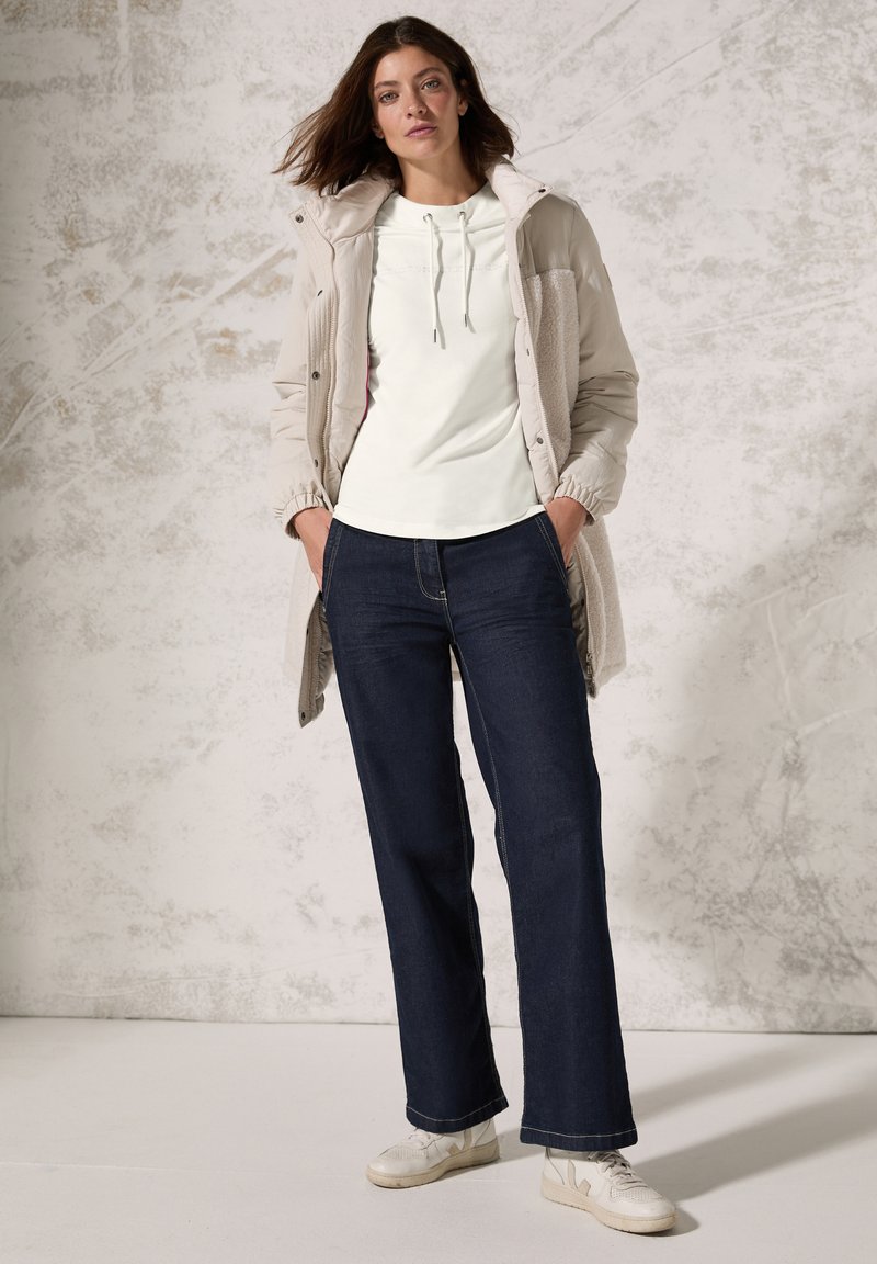 Lys beige vatteret jakke med tekstureret front, hvid hættetrøje, mørkeblå bredbensjeans og hvide sneakers, stående mod en neutral baggrund.
