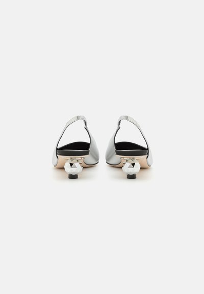 Zapatos de tacón plateados con un diseño abierto en la parte trasera y un tacón decorativo geométrico. Textura suave, con un interior negro y detalles en la base del tacón.
