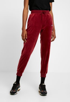Person trägt burgunderrote Velours-Jogginghose mit Kordelzugbund, schwarze Socken, bunte schwarze Sneakers und ein schwarzes Kurzarmoberteil, steht auf weißem Boden.