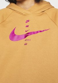 Hnědá mikina s kapucí s texturovaným povrchem. Obsahuje fialové logo Nike Swoosh a text "Just Do It." Prošité detaily podél švů.