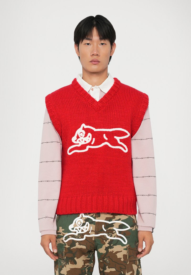 Chaleco de punto rojo con escote en V, decorado con un diseño de un gato blanco. Lleva encima una camisa de manga larga rosa claro con patrones horizontales.