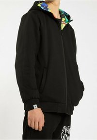Homme portant un sweat à capuche noir zippé avec une doublure intérieure colorée et un pantalon noir avec un texte blanc sur la cuisse.
