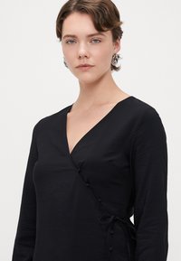 Melna aukliņu kleita ar garām piedurknēm, teksturēta auduma, V veida dekoltē, dekoratīvas pogas sānos un saistīta vidukļa detaļa.