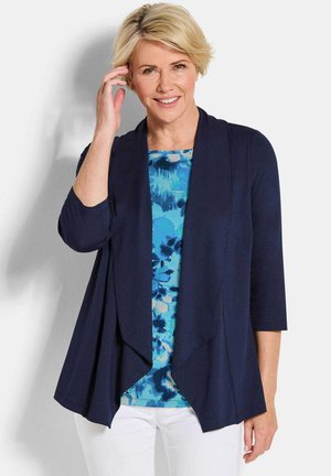 Vrouw met kort blond haar die glimlacht, draagt een marineblauwe open voorkant cardigan over een blauw patroon top en witte broek, poseert tegen een witte achtergrond.