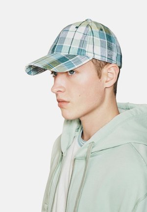 Junger Mann in mintgrünem Kapuzenpullover, der eine blau-grüne karierte Baseballkappe trägt und mit neutralem Ausdruck nach links schaut.