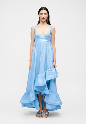 Kobieta z długimi ciemnymi włosami, ubrana w jasnoniebieską sukienkę bez rękawów z falbankami na asymetryjnym wykończeniu i sandałami z odkrytymi palcami na jednolitym tle.