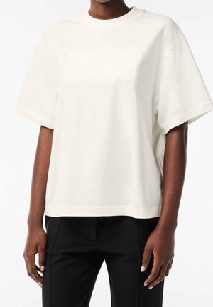Personne portant un t-shirt blanc ample à manches courtes avec un texte subtilement embossé sur la poitrine et un pantalon noir, debout sur fond blanc.