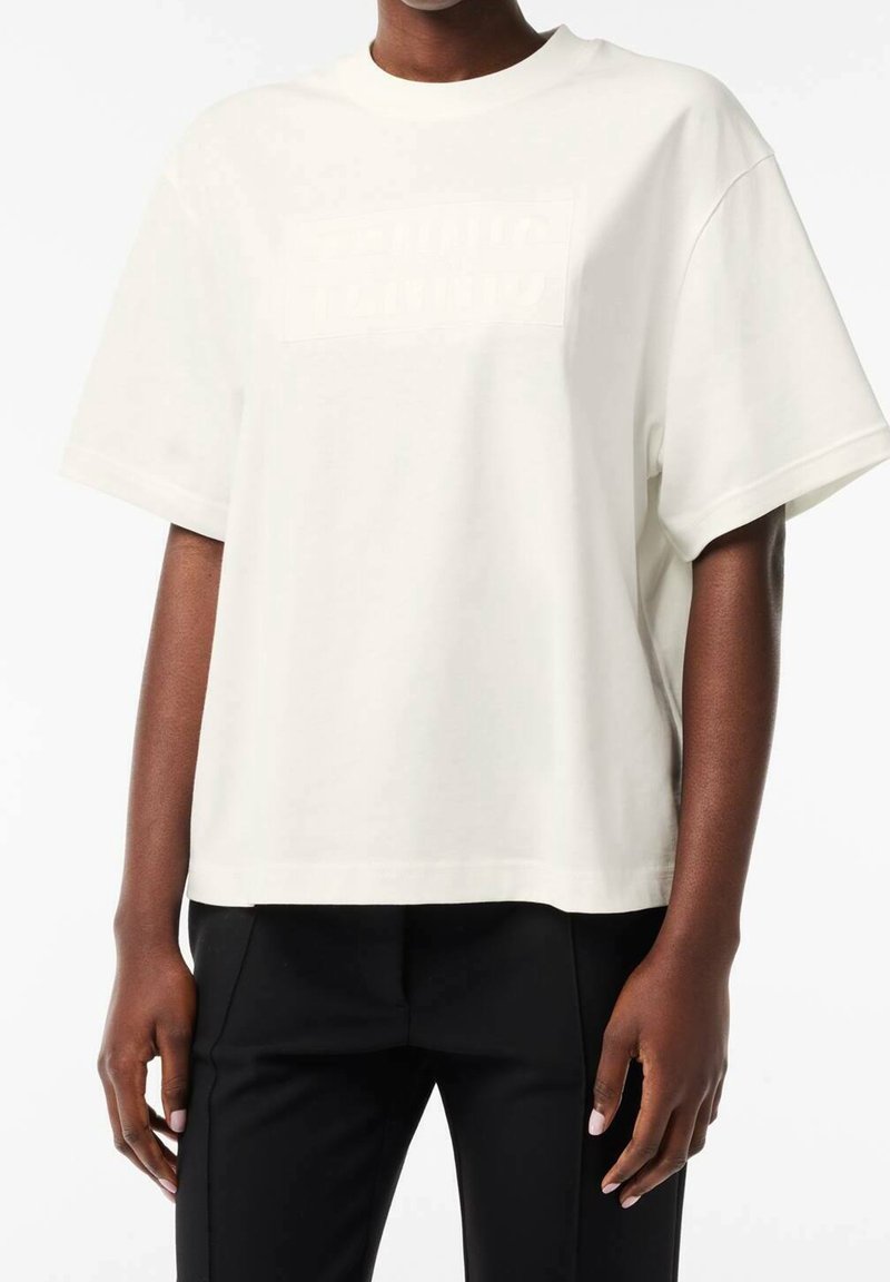 Personne portant un t-shirt blanc ample à manches courtes avec un texte subtilement embossé sur la poitrine et un pantalon noir, debout sur fond blanc.