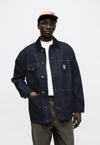 Muž s brýlemi, béžovou čepicí, tmavě modrou džínovou bundou s náprsními kapsami, bílou košilí a volnými šedými kalhotami, stojící před jednobarevným pozadím.