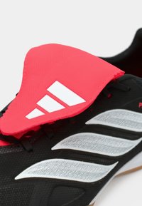 Μαύρο αθλητικό παπούτσι με ροζ λαστιχένια γλώσσα που διαθέτει λευκές ρίγες, υφή στο πάνω μέρος και ανοιχτογκρι πλευρικές λεπτομέρειες. Ορατός σχεδιασμός λαστιχένιας σόλας.