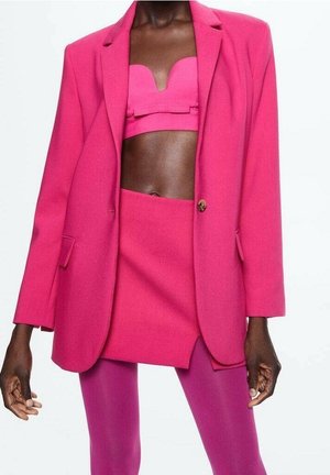 Osoba o ciemnej karnacji ubrana w jasnoróżową marynarkę, dopasowany top odsłaniający brzuch, mini spódniczkę i różowe rajstopy na jednolitym białym tle.