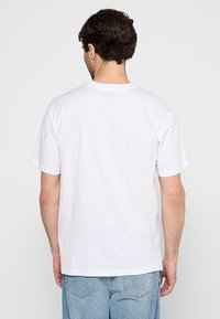 Camiseta de algodón blanca con un ajuste relajado, mangas cortas y una textura suave. La parte posterior presenta un pequeño logo cerca del escote.