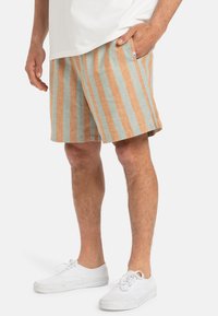 Pantaloni corti in lino con righe verticali arancioni e azzurri. Presentano una tasca laterale e una vestibilità comoda. Indossati con sneakers bianche.