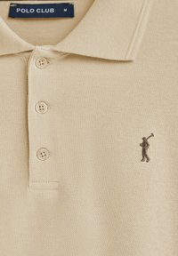 Beżowa koszulka polo z teksturowanej tkaniny, z trzema guzikami i małym brązowym haftowanym logo golfisty po lewej stronie klatki piersiowej.