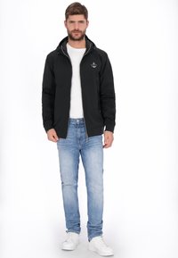 Sort lynlåsjakke med hætte, med logo på brystet, kombineret med lyseblå jeans og hvide sneakers. Glat stoftekstur.