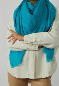 Personne portant une veste beige et un pantalon marron clair avec une écharpe bleu vif à motifs enroulée autour du cou et les bras croisés.