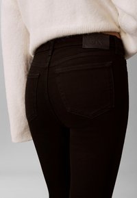 Melnas džinsa bikses ar vidēju jostas augstumu, priekšējām un aizmugurējām kabatām, un teksturētu etiķeti pie jostasvietas. Mīksts, elastīgs audums.