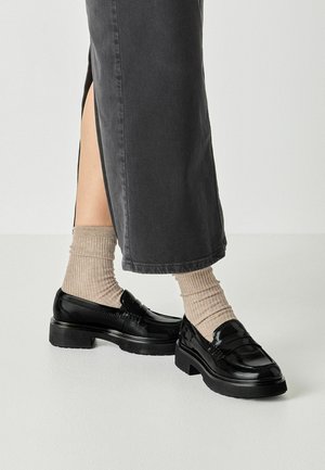 Schwarze Plateau-Loafers getragen mit beigen, gerippten Socken und einem dunkelgrauen Rock mit seitlichem Schlitz vor einfachem hellem Hintergrund.