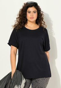T-shirt nero a maniche corte con una vestibilità rilassata, caratterizzato da dettagli con maniche arricciate. Abbinato a leggings con fantasia e una giacca grigia.