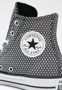 Sneakers Converse alte bianche e nere con un motivo a rete, occhielli in metallo e un logo circolare con una stella e dettagli del marchio.