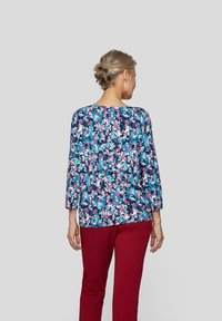 Langärmlige Bluse mit Blumenmuster in Blau mit rosa und weißen Akzenten, kombiniert mit roten Hosen. Die Bluse hat einen runden Halsausschnitt und einen lässigen Schnitt.