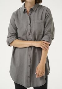 Blouse grise à manches longues en tissu doux, avec une patte de boutonnage à l'avant, une poche poitrine unique et des poignets de manches retroussés. Ourlet arrondi.