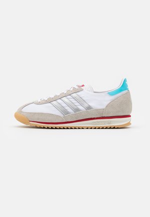 Baskets Adidas blanches et grises avec languette talon bleu clair, bande rouge sur la semelle intermédiaire, semelle extérieure en gomme et trois bandes grises sur le côté.