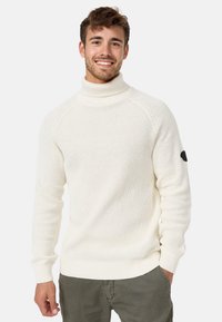 Pull en maille blanc à col roulé avec des poignets et un ourlet côtelés, présentant un motif texturé et un patch noir sur la manche gauche.