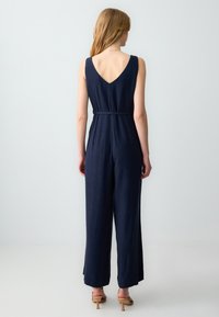 Salopette blu navy con schiena a V, design senza maniche, vita cinchata, gamba larga e texture morbida. Abbinata a tacchi con cinturini marroni.