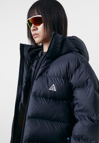 Black puffy jacket featuring a high collar and a silver triangular logo. Smooth, shiny texture with a zip closure and tightly packed insulation.