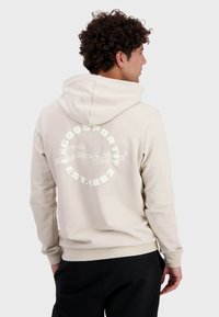 Sudadera con capucha beige con el logotipo en texto "COQ SPORTIF" y "EST. 1882" impreso en la parte posterior. Hecha de tela suave, cuenta con un ajuste relajado.