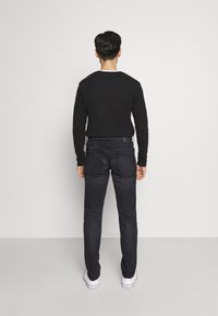 Camisa de manga larga negra, jeans de mezclilla oscuros, diseño sencillo, forma ajustada, textura sutil, complementada con zapatillas blancas sobre un fondo gris.