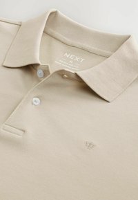 Beige poolopaita, valmistettu teksturoidusta kankaasta. Siinä on klassinen kaulus, kolmen napin läppä ja pieni kirjailtu kruunu-logo.