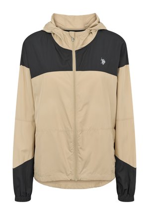 Person med armene over kors iført en beige og sort lynlåset hættejakke og sorte tætsiddende bukser mod en ensfarvet lys baggrund.