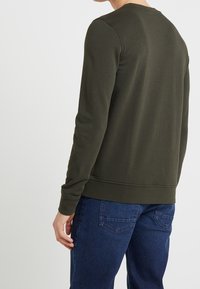 Felpa verde scuro con maniche lunghe, scollo rotondo e orlo a coste. Indossata con jeans blu; il tessuto appare liscio e informale.