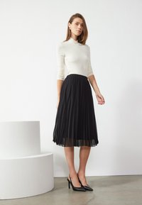 Svart plissert skjørt med gjennomsiktig overlay, kombinert med en beige langermet topp og svarte slingback-heler, vist mot en ensfarget bakgrunn.