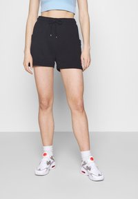 Person klädd i svarta shorts med dragsko, vita strumpor i ankellängd och vita sportskor med röda detaljer, stående på ett ljusgrått golv.