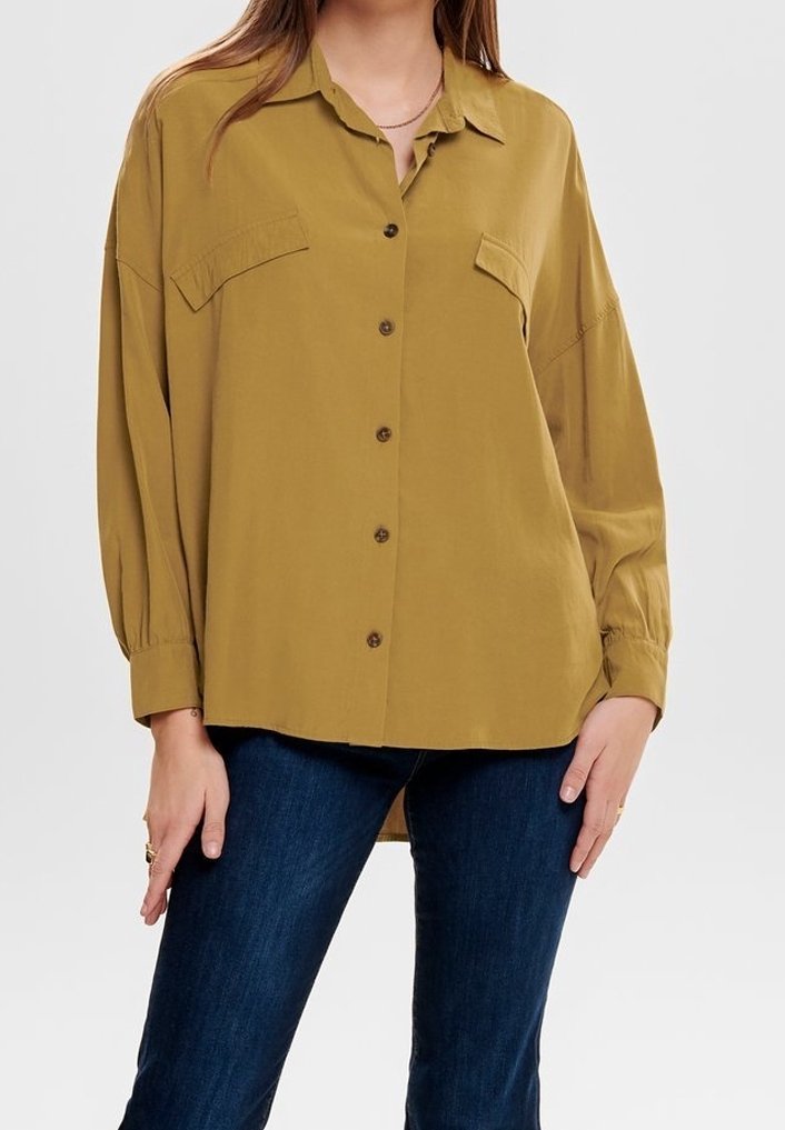 Femme portant une chemise boutonnée oversize jaune moutarde avec des poches poitrine et un jean bleu foncé sur un fond uni.