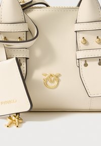 Bézs bőrtáska aranyszínű kiegészítőkkel, díszítő varrással és két felső fogantyúval. Tartozéka egy levehető kulcstartó táska.