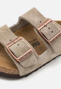 Sandały Birkenstock w jasnoszarym zamszu z dwoma regulowanymi klamrami w kolorze miedzi, z korkowym wkładką i teksturowaną brązową podeszwą.
